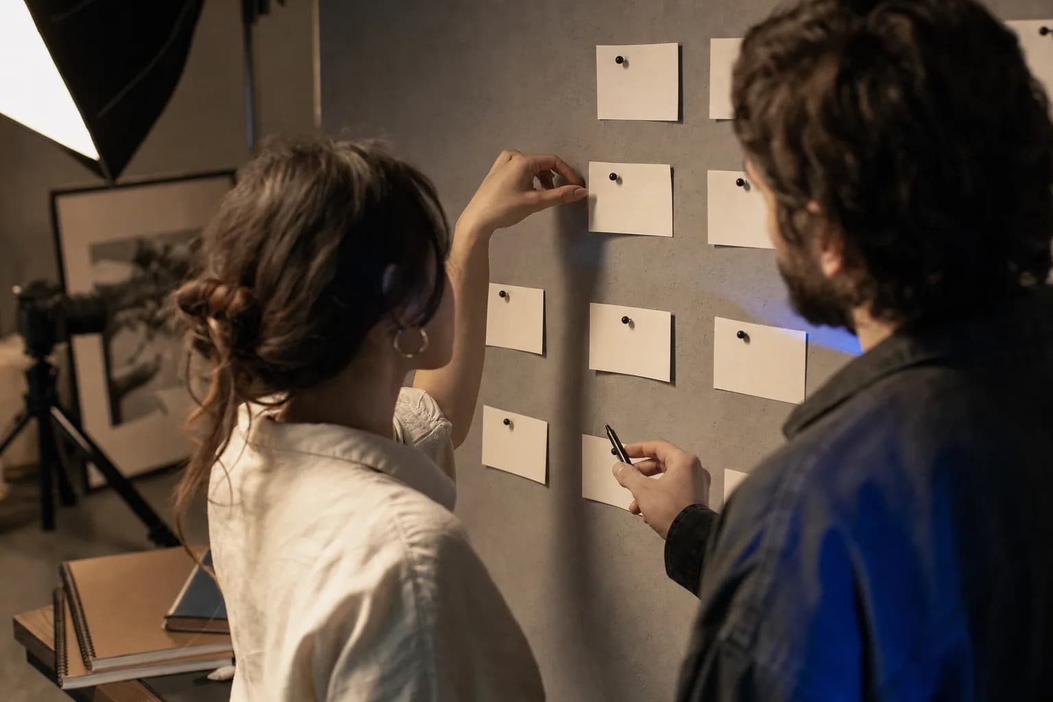 Two creative strategists in a modern studio examining a single index card pinned to a wall of blank cards, lit by warm directional light with a cool blue rim from a nearby monitor.